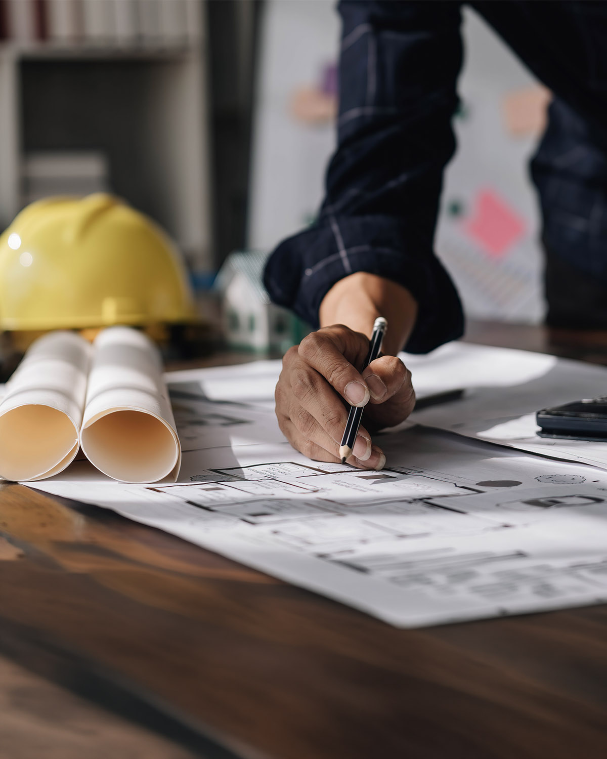 Hände eines Ingenieurs auf einem Konstruktionsplan, Bleistift, gelber Bauhelm und aufgerollte Pläne auf Holztisch