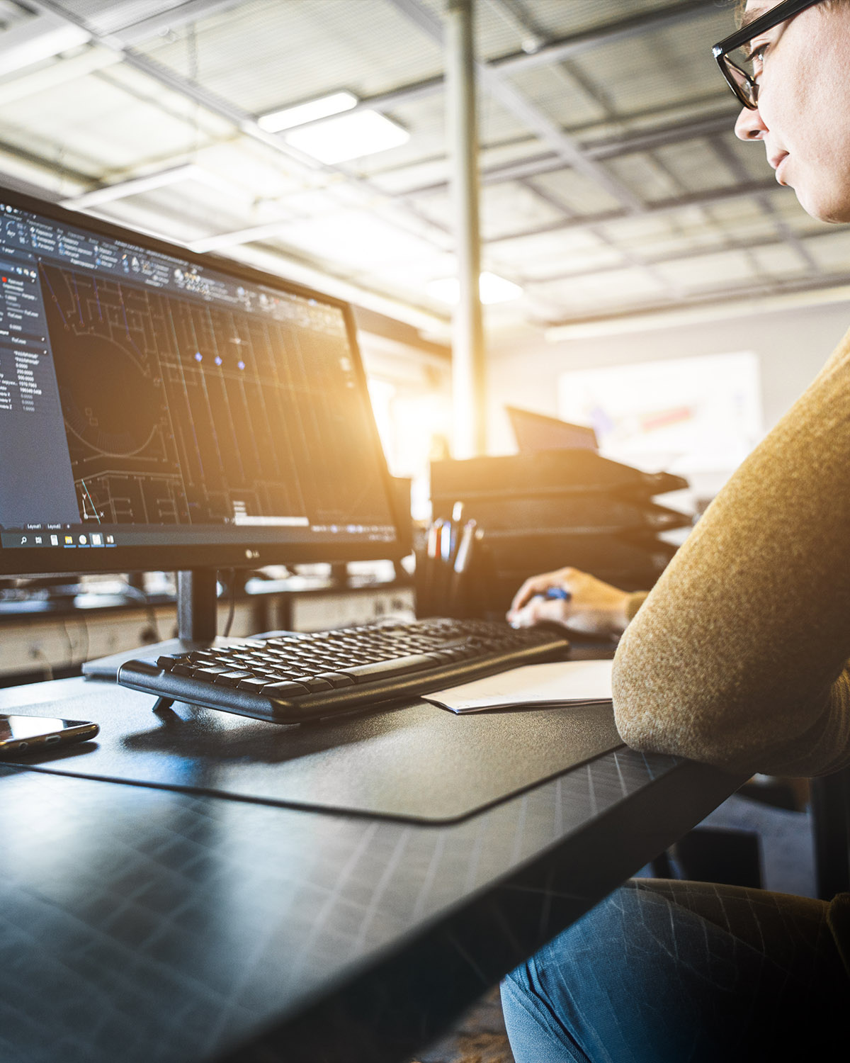 Ingenieurin am Schreibtisch vor großem Monitor mit AutoCAD-Zeichnung, Industrie-Halle im Hintergrund
